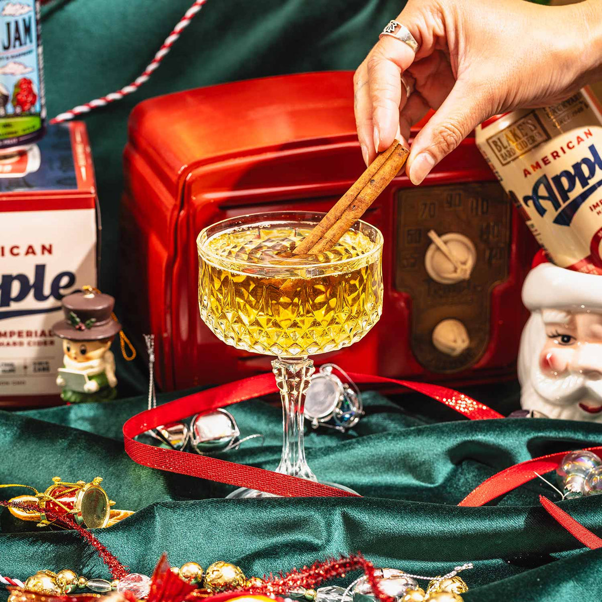 An American Pie cocktail garnished with a cinnamon stick, with cans of Blake's Hard Cider American Apple