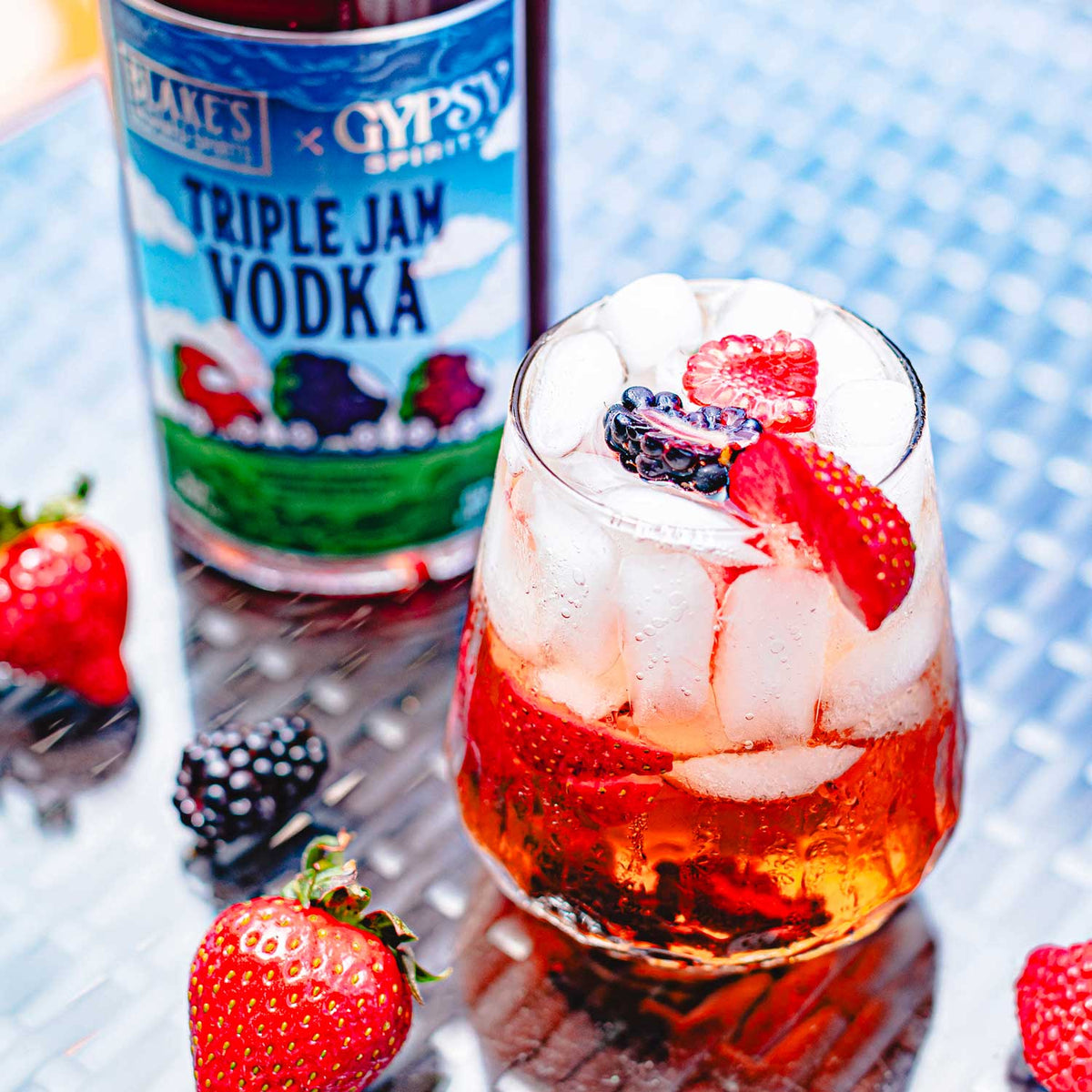 A Triple Berry Vodka Soda garnished with fresh berries, beside a bottle of Blake's x Gypsy Spirits Triple Jam Vodka