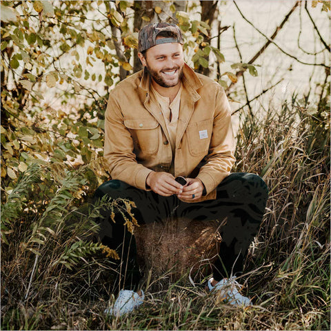 A man in a canvas jacket sitting in tall grass on an autumn day